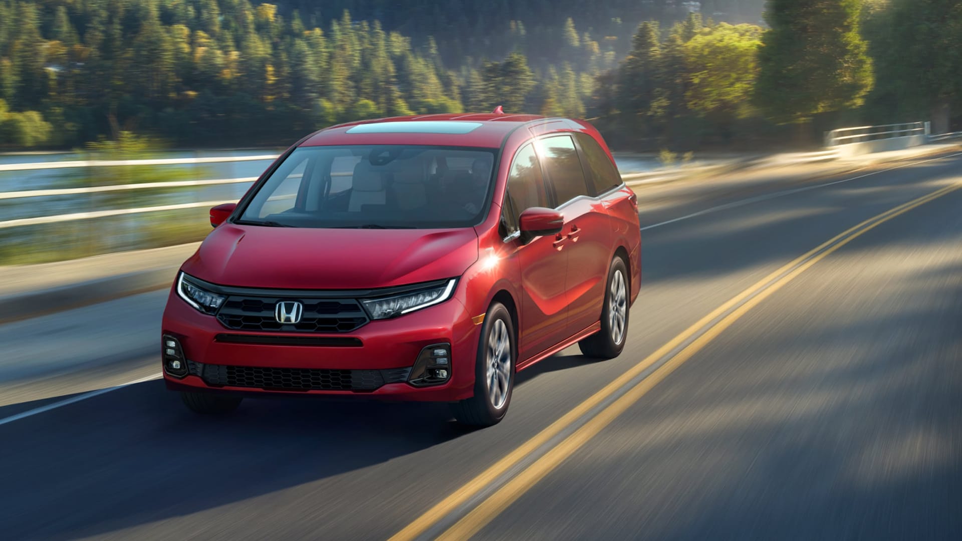 A red Honda Odyssey driving down the road by a lake and a forest
